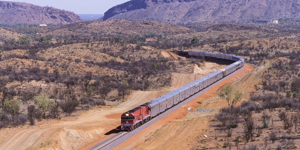 "Alice Springs, Australia - February 2, 2004: The first Ghan passenger train to Darwin departs Alice Springs with the spectacular MacDonnell Ranges in the background.  The extra long VIP train is mid-way through its inaugural 2969km transcontinental journey from Adelaide in the South to Darwin in the North.  The first train departed Adelaide February 1 and arrived in Darwin to a huge welcome on February 3, 2004.  The journey from the Mediterranean South through Australias Red Centre to the tropical Top End takes the Ghan 54hrs.  The Ghans symbol is a camel and its handler, in recognition of the pioneering Afghan cameleers who opened up the outback of Australia in the mid-19th century."